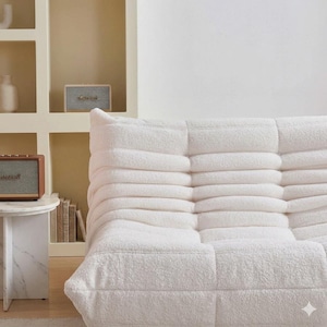 May include: A white, plush, modular sofa with a textured, tufted design. The sofa is the focal point of the image, with a small marble side table and a vintage-style speaker visible in the background. The room has a neutral color palette.