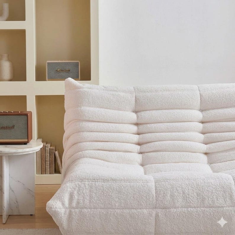 May include: A white, plush, modular sofa with a textured, tufted design. The sofa is the focal point of the image, with a small marble side table and a vintage-style speaker visible in the background. The room has a neutral color palette.