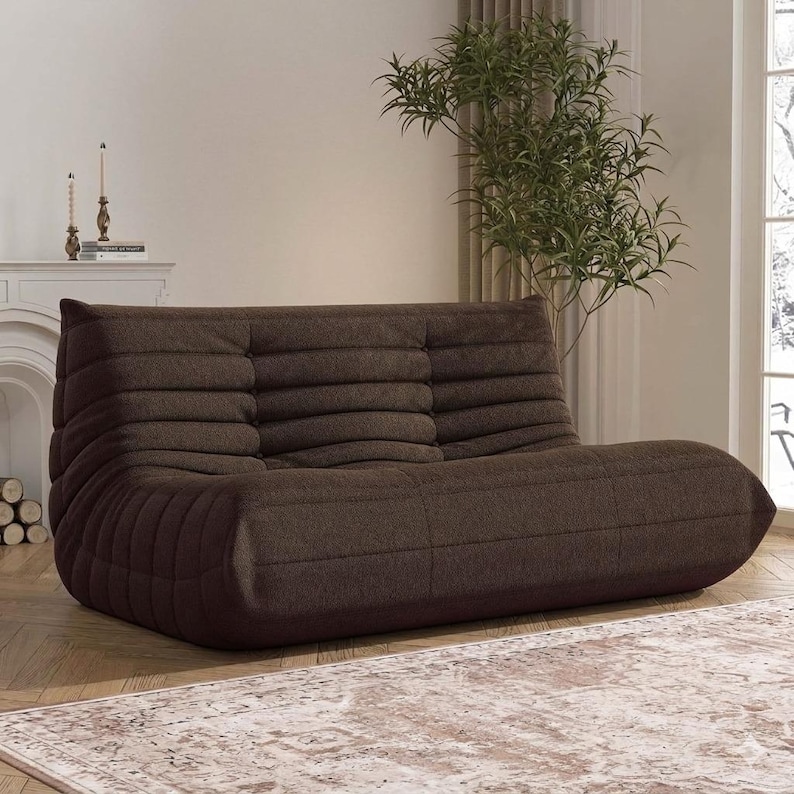May include: A brown, low-profile sofa with a textured, ribbed design. The sofa has a rounded shape and is placed in a well-lit room with a rug and a plant. The sofa is a modern piece of furniture.