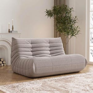 May include: A light gray, low-profile sofa with a quilted design. The sofa has a rounded base and a backrest with horizontal stitching. The room features a fireplace, candles, and a decorative rug, creating a cozy atmosphere.