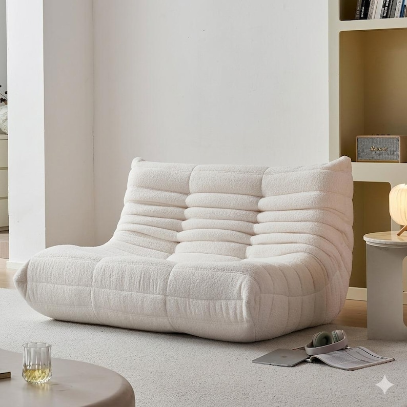 May include: A cream-colored, low-profile sofa with a textured, ribbed design. The sofa has a rounded shape and is made of a soft, plush material. The sofa is placed on a light-colored carpet in a well-lit room.