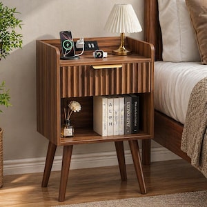 May include: A wooden nightstand with a drawer and open shelf. The nightstand has a lamp, a phone charging station, and a small vase with a flower. The drawer has a gold handle. Books are on the shelf. The nightstand is next to a bed.