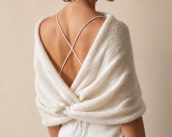 May include: A cream-colored, soft-knit wrap featuring a twisted back design and long sleeves that drape over the shoulders. The garment is worn over a white dress with thin, crisscrossed spaghetti straps that are visible against the back.
