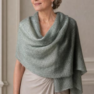 Puede incluir: Un chal tejido de color verde salvia con una textura suave y difusa, drapeado sobre los hombros. La prenda presenta una construcción de punto suelto que crea una apariencia ligera y aireada, ofreciendo una capa delicada sobre un vestido beige.