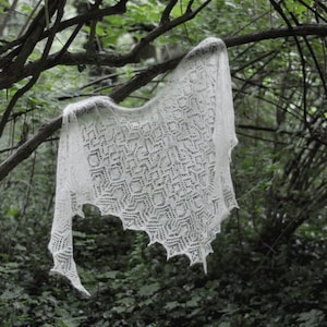 May include: A white crocheted shawl with a geometric pattern hangs from a tree branch in a forest setting.