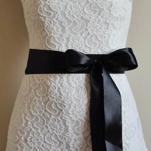 Puede incluir: Un vestido blanco de encaje con una cinta de satén negro atada en un lazo en la cintura.