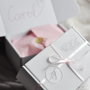 May include: A white cardboard gift box tied with a thin pink ribbon and a circular sticker featuring the letters WF. The box lid displays a grey floral illustration. Inside, pink tissue paper is sealed with a gold wax stamp, and the name Carol is written in pink script on the inner lid.