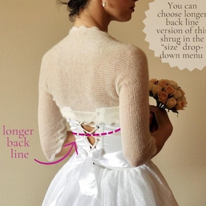 May include: A cream-colored knit shrug with three-quarter length sleeves worn over a white satin dress featuring a lace-up back closure with a ribbon tie. Text on the image reads: You can choose longer back line version of this shrug in the size drop-down menu and longer back line.