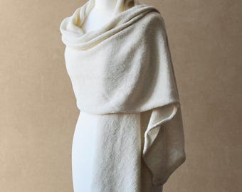Puede incluir: Un chal de punto color crema drapeado sobre un maniquí. El tejido presenta una construcción de punto fino y ligero con una textura suave y una apariencia sutilmente semitransparente. La tela cae en pliegues sueltos alrededor del cuello.