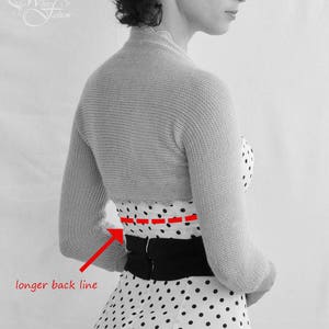 Puede incluir: Una foto en blanco y negro de una mujer que lleva un vestido blanco de lunares con un cintur&oacute;n negro. Lleva una chaqueta de punto gris con una l&iacute;nea de espalda m&aacute;s larga.
