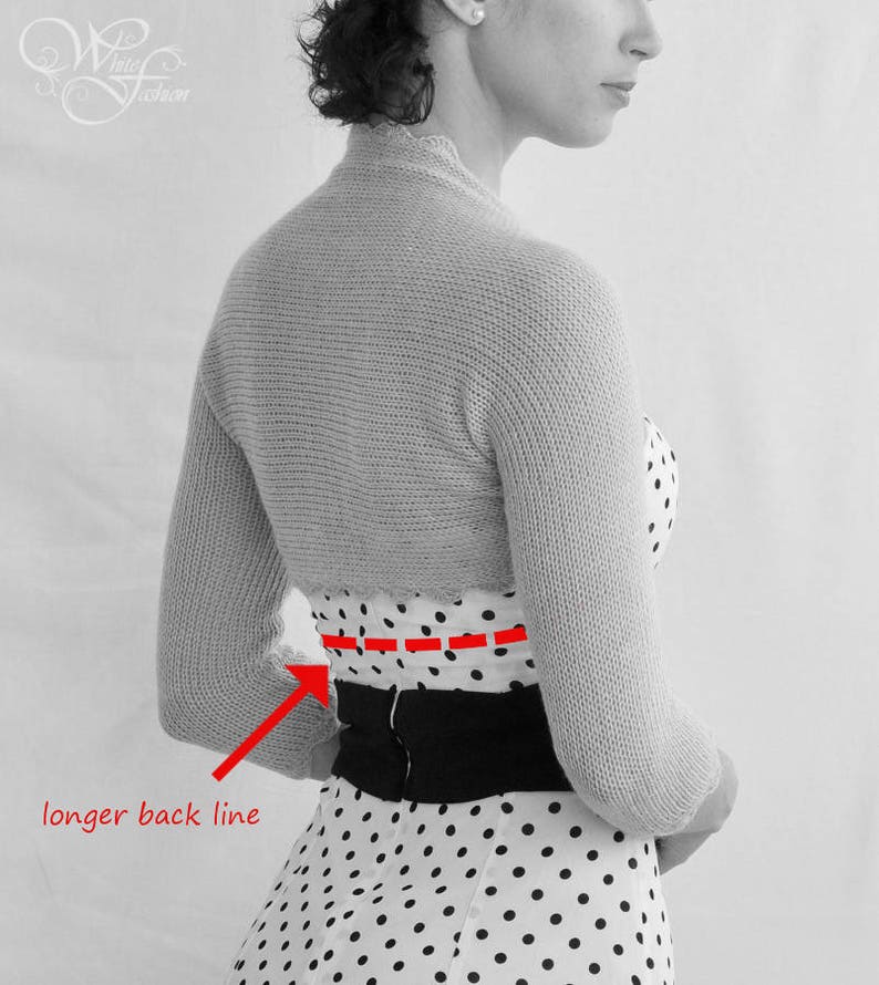 Puede incluir: Una foto en blanco y negro de una mujer que lleva un vestido blanco de lunares con un cintur&oacute;n negro. Lleva una chaqueta de punto gris con una l&iacute;nea de espalda m&aacute;s larga.
