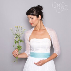 May include: A white sheer knit bolero with three-quarter length sleeves worn over a strapless white dress featuring a light blue waistband. The garment has a delicate, semi-transparent texture and a scalloped edge along the front opening.