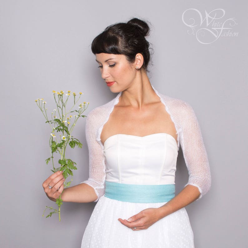 May include: A white sheer knit bolero with three-quarter length sleeves worn over a strapless white dress featuring a light blue waistband. The garment has a delicate, semi-transparent texture and a scalloped edge along the front opening.