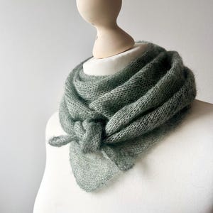 Puede incluir: Una bufanda de punto a mano, de color verde claro, está drapeada alrededor del cuello de un maniquí. La bufanda está anudada en la parte delantera, creando un efecto texturizado y en capas. El tejido suave y aireado sugiere un accesorio ligero y cómodo.