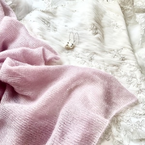 May include: A light pink, open-weave shawl draped over a white, embroidered wedding dress. Delicate pearl earrings with silver hooks rest on the dress. The shawl's texture contrasts with the smooth fabric of the dress.