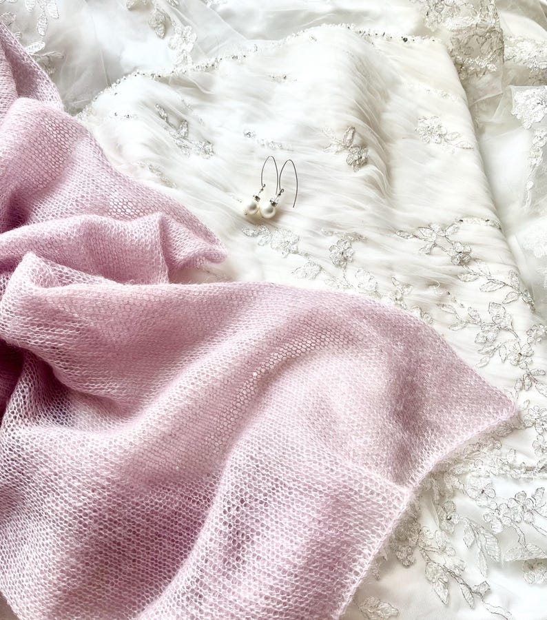 May include: A light pink, open-weave shawl draped over a white, embroidered wedding dress. Delicate pearl earrings with silver hooks rest on the dress. The shawl's texture contrasts with the smooth fabric of the dress.