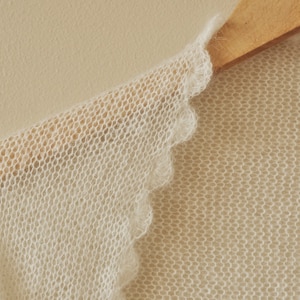 May include: A sheer, off-white knitted garment featuring a delicate, scalloped edge detail. The fabric has a soft, fuzzy texture with a loose, open-knit construction, showcasing a light, airy appearance and a consistent, fine-gauge stitch pattern.