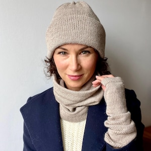 Può includere: Un set di berretto, scaldacollo e guanti senza dita in maglia beige. Gli accessori in maglia a coste sono di colore neutro, adatti al clima freddo. Il set è mostrato su una persona che indossa un cappotto blu navy.