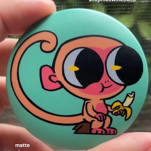 May include: A round matte pin with a teal background featuring a cartoon monkey with large black eyes, a peach-colored body, and pink accents. The monkey is holding a yellow banana. The text shopmeowmeowco is printed in white at the top right corner.