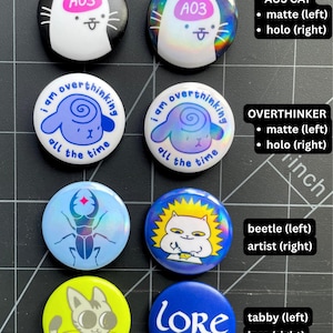 May include: Eight circular pin-back buttons arranged in two columns. Designs include a white cat with pink A03 text, a blue sheep with the text I am overthinking all the time, a blue beetle, a white cat artist, a grey tabby cat, and blue text reading LORE enthusiast. Some buttons have a matte finish while others have a holographic finish.