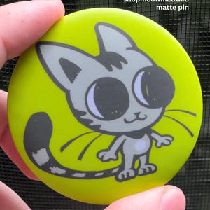 May include: A round matte pin with a bright lime green background featuring a gray cartoon cat illustration with large black eyes and a long striped tail. The text shopmeowmeowco matte pin is printed in white in the upper right corner.