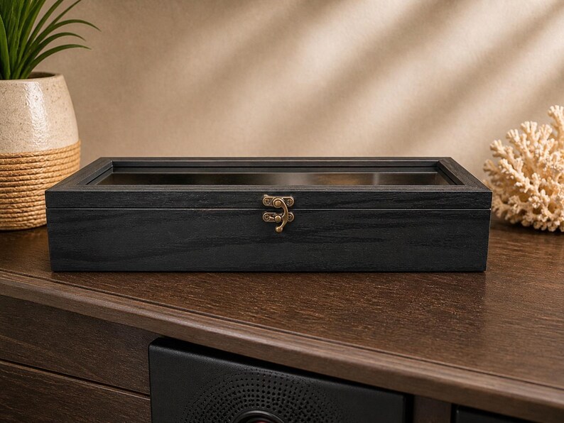 Puede incluir: Una caja de exhibici&oacute;n rectangular de madera negra con tapa de cristal y cierre color lat&oacute;n. La caja est&aacute; sobre una superficie de madera marr&oacute;n oscuro. Una planta decorativa y coral en el fondo.