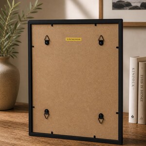 May include: A black picture frame with a brown backing, featuring four metal hanging brackets. A yellow sticker on the back reads "Do Not Hang Vertically". The frame is displayed on a wooden surface, with books and a vase visible in the background.