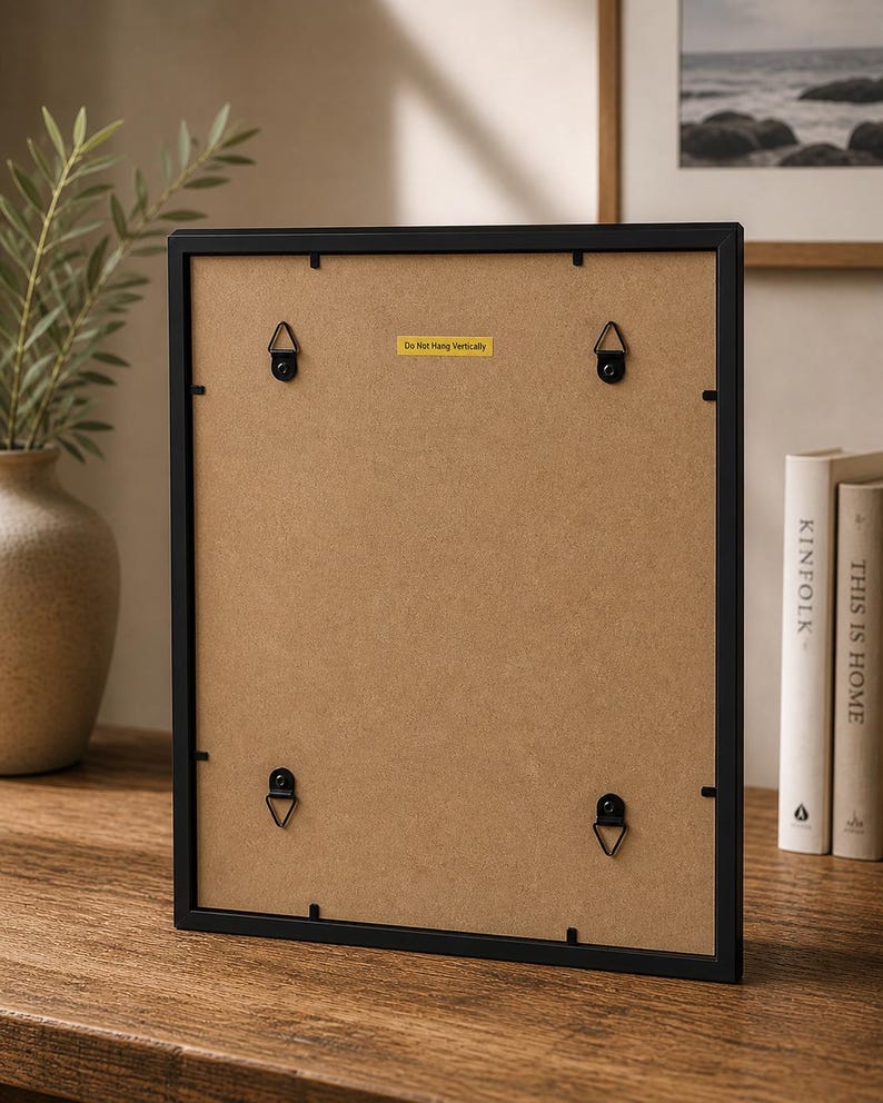 May include: A black picture frame with a brown backing, featuring four metal hanging brackets. A yellow sticker on the back reads "Do Not Hang Vertically". The frame is displayed on a wooden surface, with books and a vase visible in the background.
