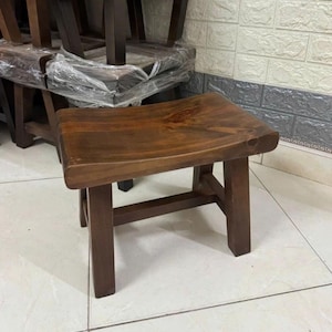 Puede incluir: Un taburete pequeño de madera marrón oscuro con un asiento rectangular curvado y cuatro patas rectas conectadas por barras de soporte horizontales. La veta de la madera es visible en la superficie lisa del asiento con un nudo natural.