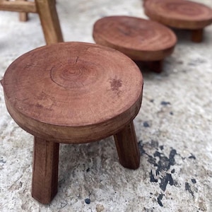 May include: A set of three small, round wooden stools with natural wood grain patterns. Each stool features a flat, circular top surface and three sturdy, straight legs, crafted from a dark brown, solid wood material with a smooth finish.