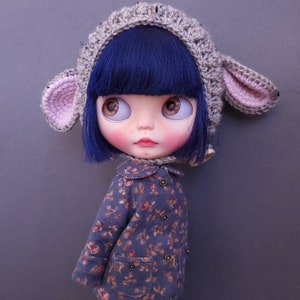 May include: A doll with blue hair wearing a gray crocheted hat with pink ears and a blue floral print jacket.