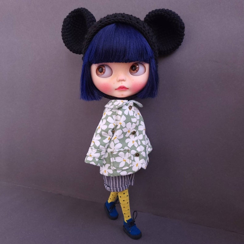 May include: A doll with black ears, a black and white hat, a green and white floral jacket, and blue shoes. The doll has blue eyes and black hair.