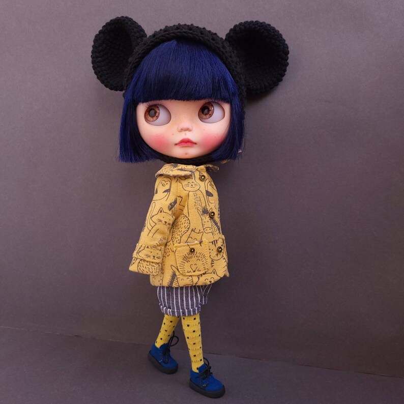 May include: A doll with blue hair wearing a yellow jacket with a cat print, black and white striped pants, yellow polka dot tights, and blue shoes. The doll is wearing a black hat with ears.