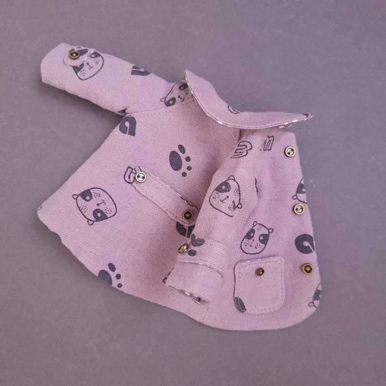 May include: A pink baby jacket with a panda print and gold buttons. The jacket has a small pocket on the front.