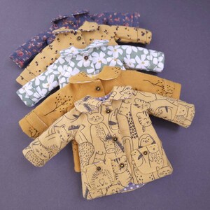 May include: A stack of four miniature doll coats in various patterns. The top coat is a dark blue with white floral print, the second is a yellow with white polka dots, the third is a green with white flowers, and the bottom coat is a yellow with a black animal print.