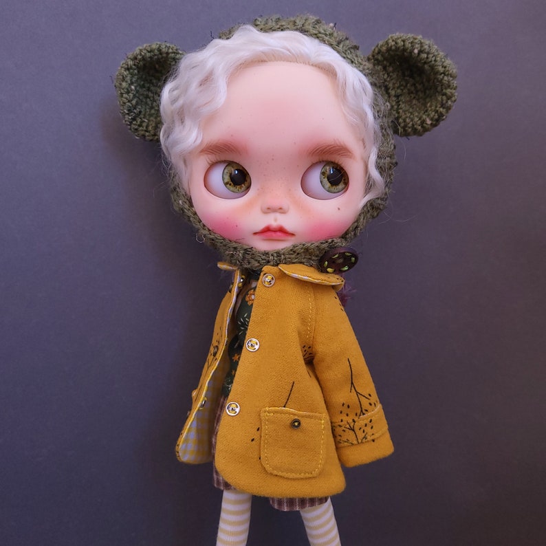May include: A doll with blonde hair wearing a green knitted hat with bear ears and a yellow jacket with brown trim. The doll is wearing a plaid skirt and white and brown striped tights.