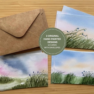 Peut inclure: Un ensemble de 12 cartes de vœux avec enveloppes présentant trois motifs originaux peints à la main à l'aquarelle de champs herbeux sous des ciels bleus et roses. Le texte sur l'incrustation circulaire indique 3 ORIGINAL HAND-PAINTED DESIGNS 12 CARDS WITH ENVELOPES.