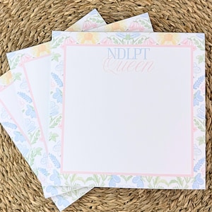May include: Stack of floral-bordered notepads with a pink border and the text "NDLPT Queen" in light blue and pink. The notepads feature a pastel floral design in blue, yellow, and pink, with a white interior.