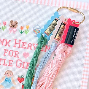 May include: Three plastic thread bobbins in light blue, white, and pink, held together by a gold-toned metal ring. Each bobbin holds a skein of embroidery floss in green, light blue, and pink. The floss is labelled Pepper Pot Silk 100% Silk 30yds.