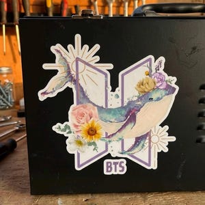 May include: A sticker featuring a watercolor-style whale with floral accents, including pink roses and a yellow sunflower. The design incorporates the letters "BTS" in a purple frame, with sunburst and floral elements.