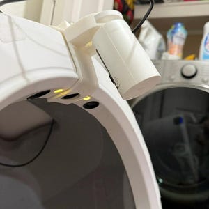 May include: A white 3D-printed plastic mounting bracket attached to the top edge of a white curved device. A white cylindrical Ring security camera is secured to the bracket, with a black power cable extending from the top of the camera unit.