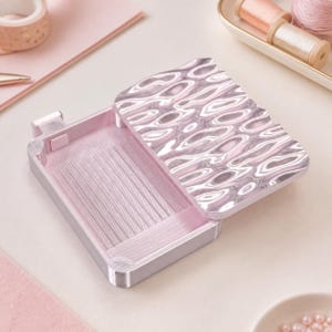 May include: A rectangular light pink needle case with a hinged lid featuring a metallic, wavy, rippled texture. The interior base is lined with a textured, ribbed surface designed to hold needles in place, with a small latch on the side.