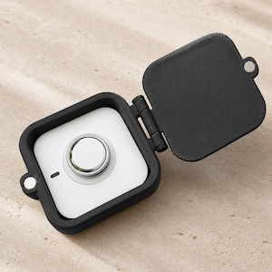 May include: A square black protective case with an open hinged lid, containing a white square electronic tracker with a central silver circular button and a small black indicator light on the front surface of the device.