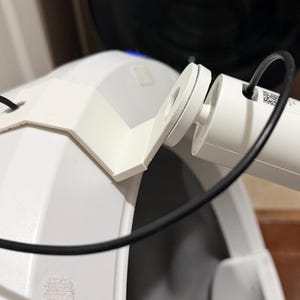 May include: A white plastic camera mount attached to a white curved surface, holding a white cylindrical security camera. A thin black power cable loops from the camera through a hole in the mount, connecting to the back of the camera unit.