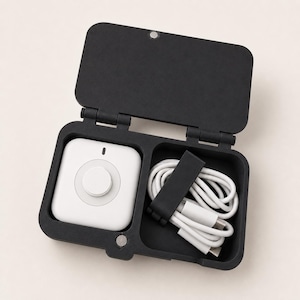 May include: A black rectangular storage case with a hinged lid containing a white square electronic device with a central circular button and a white USB-C charging cable secured by a black silicone strap within a separate compartment.