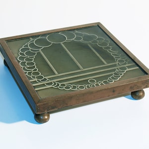 Könnte beinhalten: Quadratischer, bronzefarbener Untersetzer mit Glasplatte. Das Glas hat ein silberfarbenes, kreisförmiges Design am Rand und lineare Akzente. Der Untersetzer wird von vier kleinen, abgerundeten Füßen getragen. Ein dekorativer Artikel für das Zuhause.