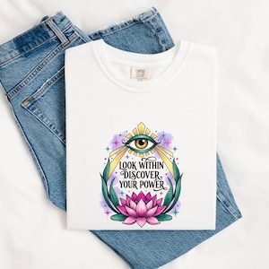 May include: A white t-shirt featuring a graphic print of a stylized eye above the text "LOOK WITHIN DISCOVER YOUR POWER" and a pink lotus flower at the base, framed by green leaves and small colorful star accents on a light purple background.