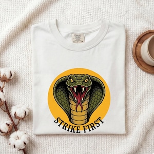 May include: A white t-shirt featuring a graphic of a green cobra snake with yellow eyes and a red tongue centered within a yellow circle. Below the snake, the text STRIKE FIRST is printed in black capital letters. The shirt has a crew neckline.