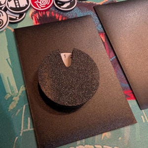 May include: A black, textured, circular game piece with a notch, resting on a black rectangular card. The number "1" is visible in the notch. Other game pieces with various designs and colors are in the background.