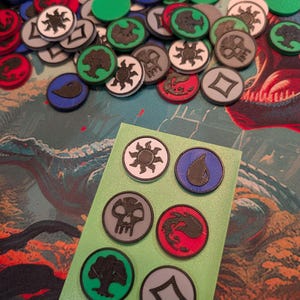 May include: A collection of colorful, circular game tokens with various symbols, including a sun, a water droplet, a skull, a dragon, a tree, and a diamond. The tokens are in shades of red, blue, green, gray, and white. Some tokens are scattered, while others are on a green card.